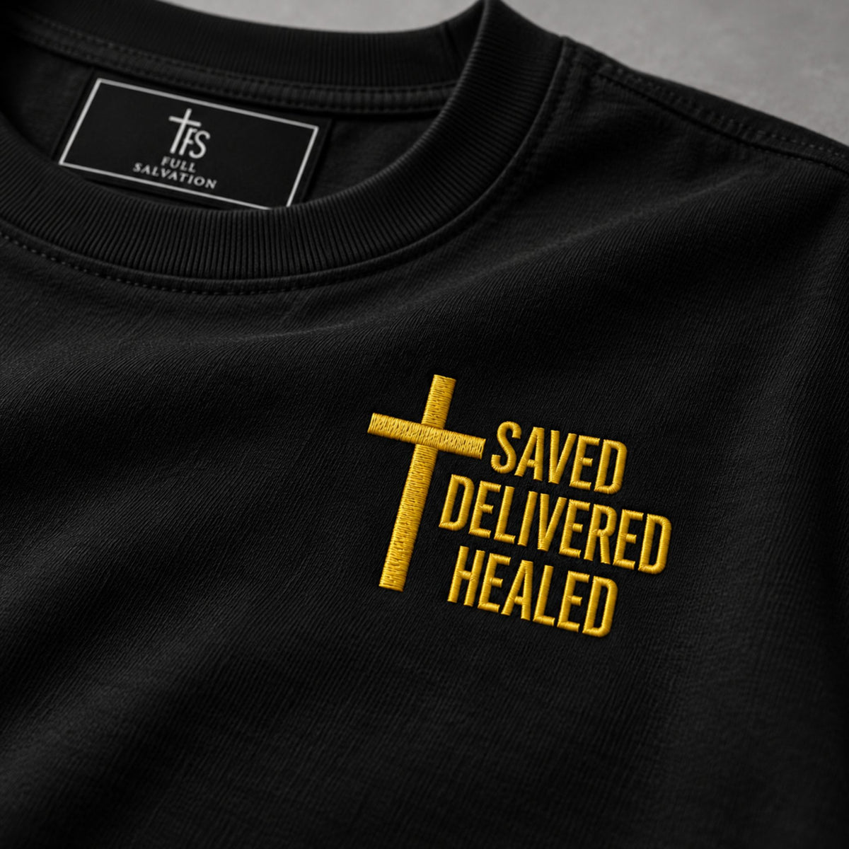 Macro detail of the gold embroidered cross and Saved Delivered Healed lettering on black cotton